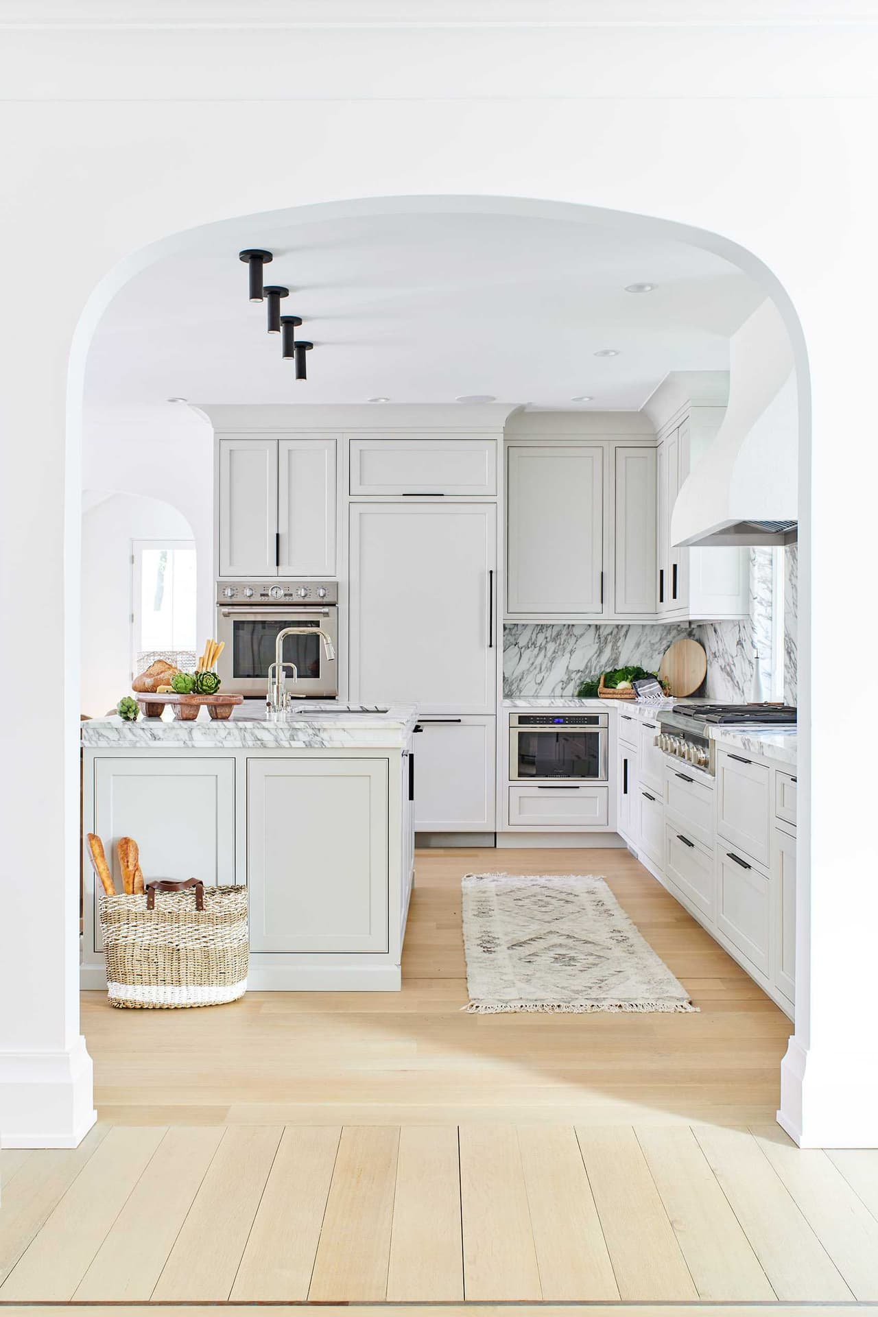 Kitchen seen through a tall arched cased opening, with pale cabinetry, a marble island and white oak floors