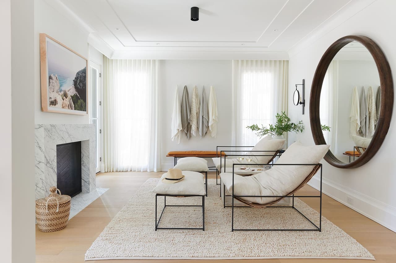 Long room in the Mroz family home in Westfield, NJ, with a marble fireplace, a leaning round mirror and a pair of sling chairs on a deep rug