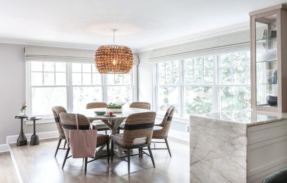 Dining area with a round table, a woven drum pendant and a bank of windows, beside a marble-topped counter
