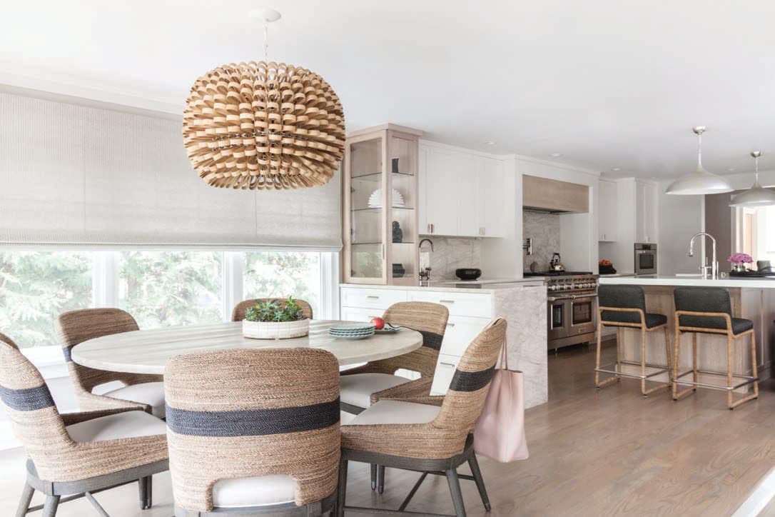 Round dining table under a woven pendant, open to a kitchen with a navy island and counter stools