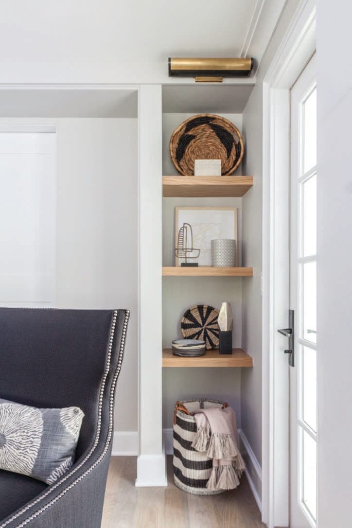 Built-in niche with oak shelves, woven baskets and a brass picture light, beside a navy armchair