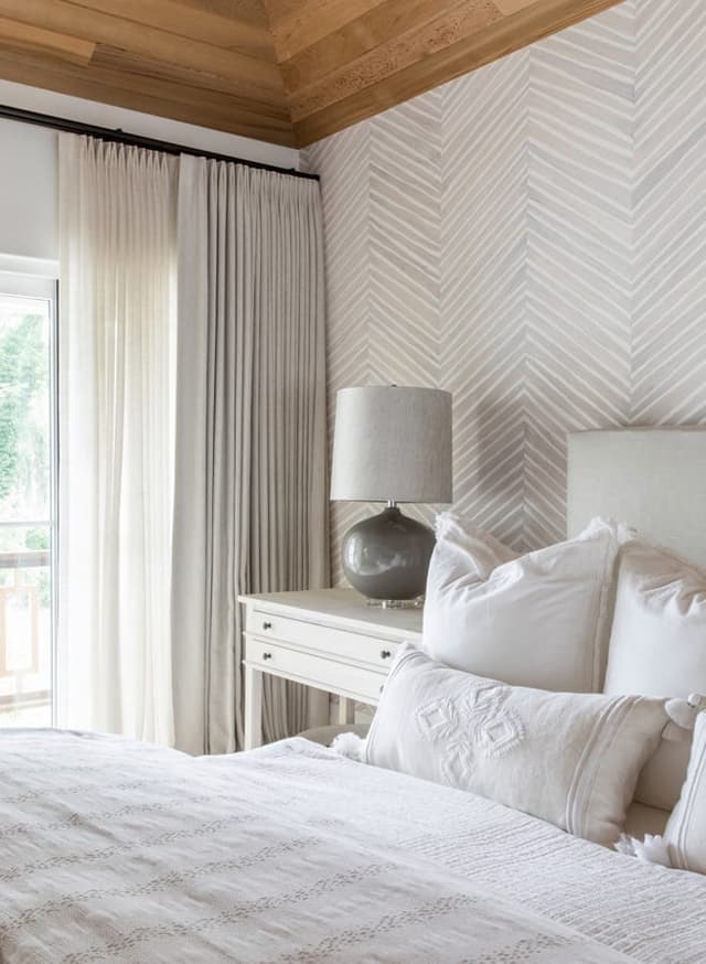 Bed against a herringbone-papered wall in a Westfield, NJ bedroom, under a planked wood ceiling with linen curtains alongside