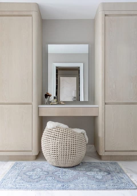 Dressing table set into pale oak cabinetry in a Westfield, NJ home, with a woven stool pushed under it on a faded blue rug