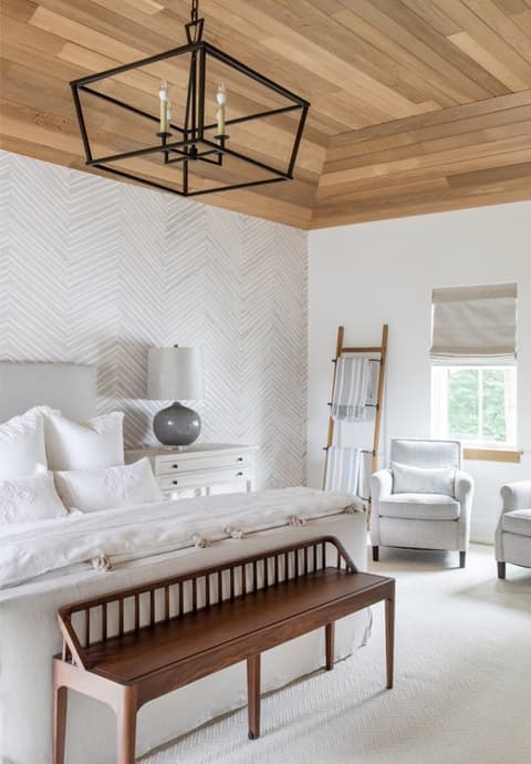 Primary bedroom of a Westfield, NJ home with a planked wood ceiling, a herringbone-papered wall behind an upholstered bed, a black lantern chandelier and a walnut spindle bench at the foot