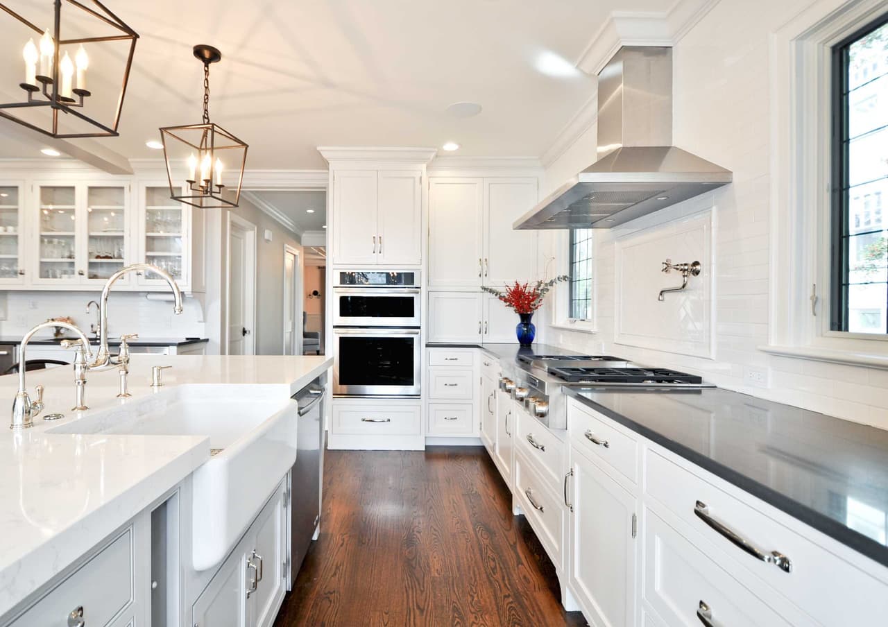 Galley kitchen with white cabinetry, a farmhouse sink, a stainless hood and two lantern pendants over the counter
