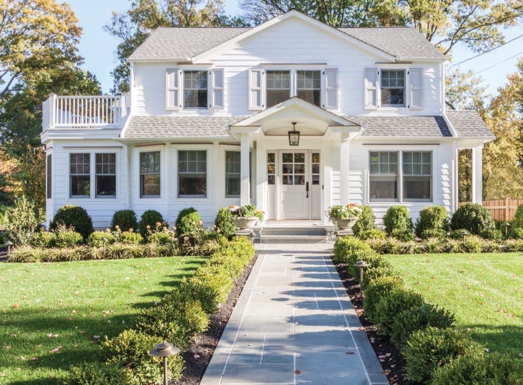Front of a pale blue shingled Westfield, NJ home, with a gabled entry porch, a roof deck behind the balustrade and a bluestone walk between clipped hedges