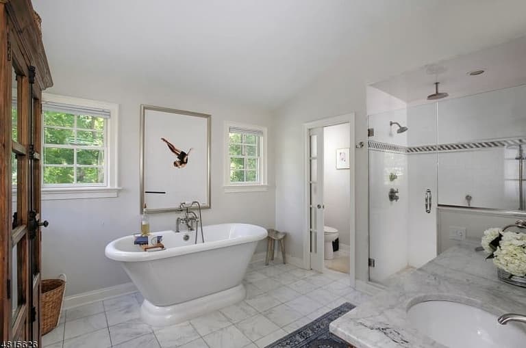 Primary bathroom of a Westfield, NJ coastal colonial renovation, with a rolltop tub under two windows and a glass shower along the marble-tiled wall