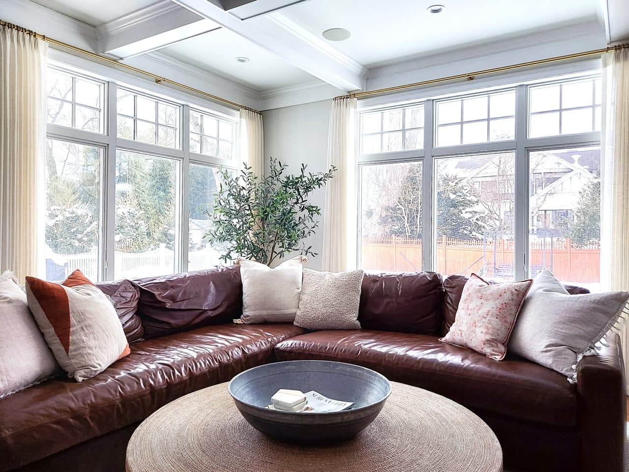 Family room of a Westfield, NJ renovation under a coffered ceiling, with a brown leather sectional in the window bay and an olive tree in the corner