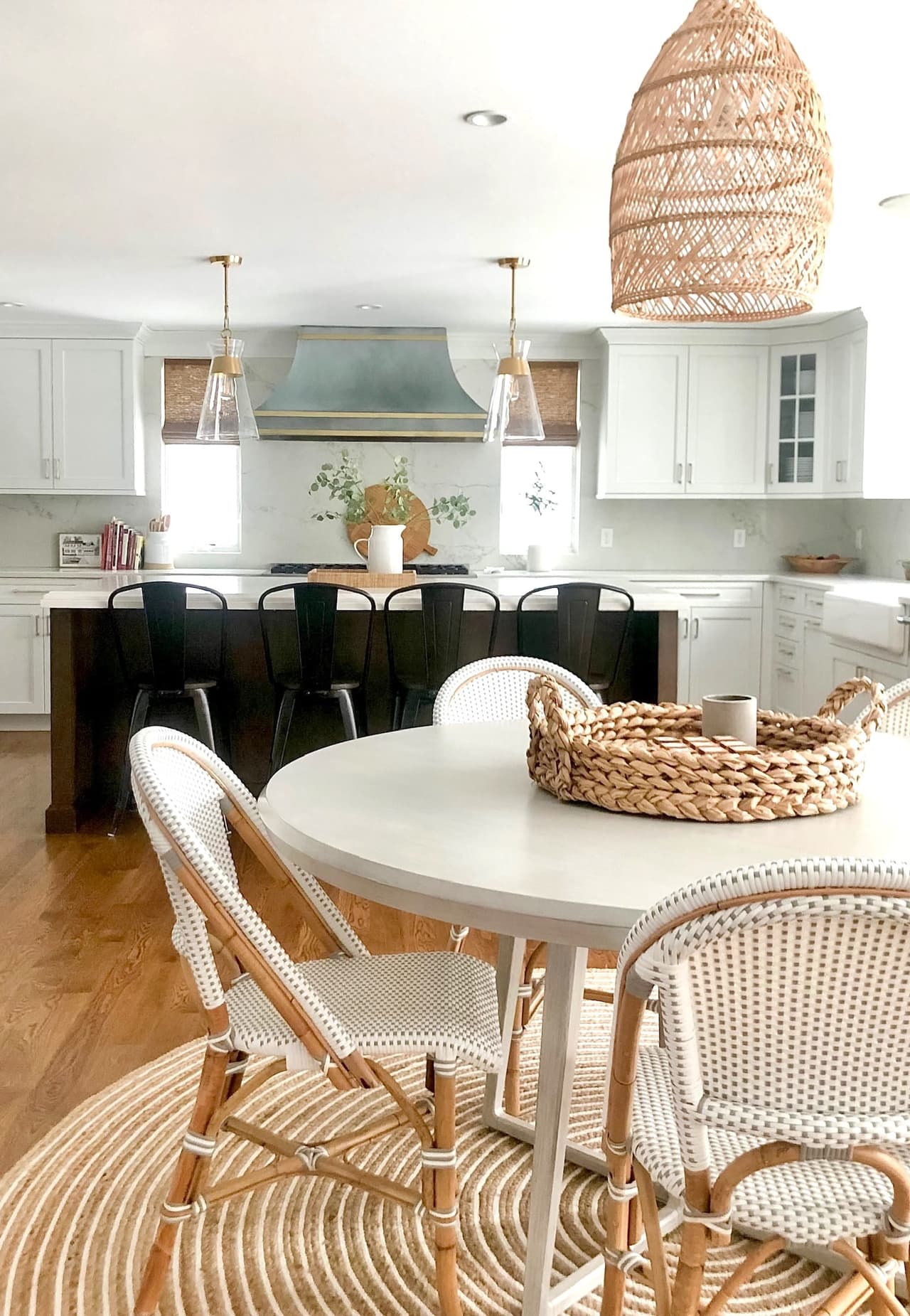 Round dining table with rattan bistro chairs under a woven pendant, open to the kitchen beyond