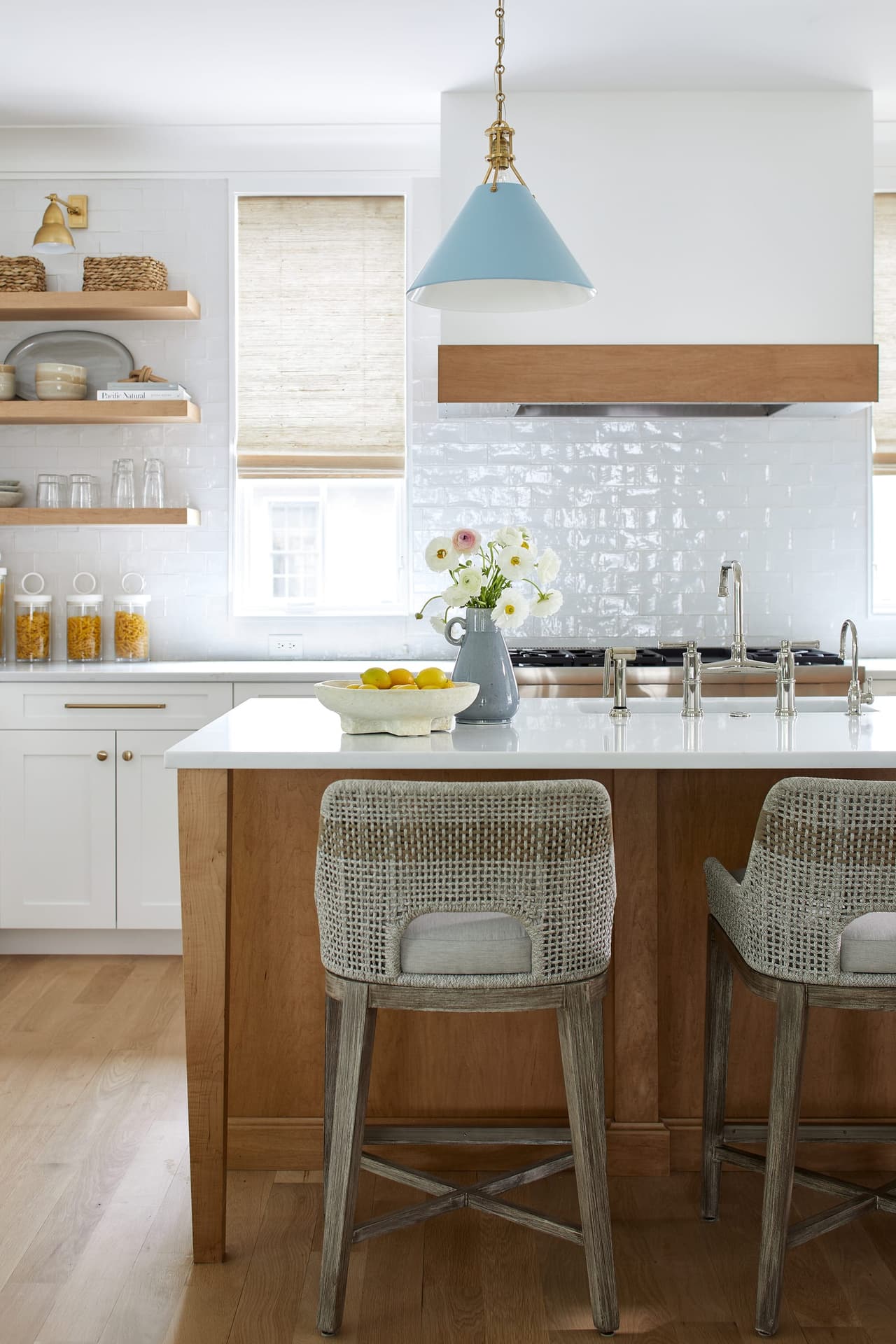Kitchen island in white oak with woven counter stools, a pale blue pendant above and glazed white tile behind the range