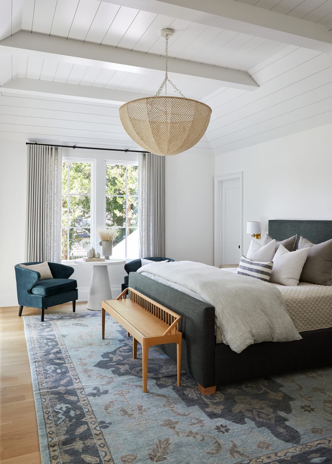 Primary bedroom under a vaulted planked ceiling, with a woven pendant and a bench at the foot of the bed