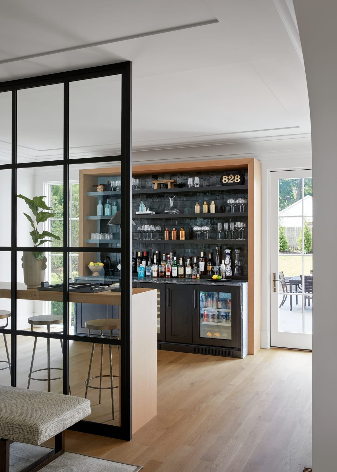 Bar behind a steel-and-glass partition, with open shelving, a beverage refrigerator and counter stools