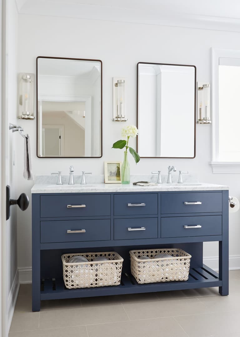 Navy double vanity with a marble top, bronze-framed mirrors, fluted glass sconces and woven baskets below