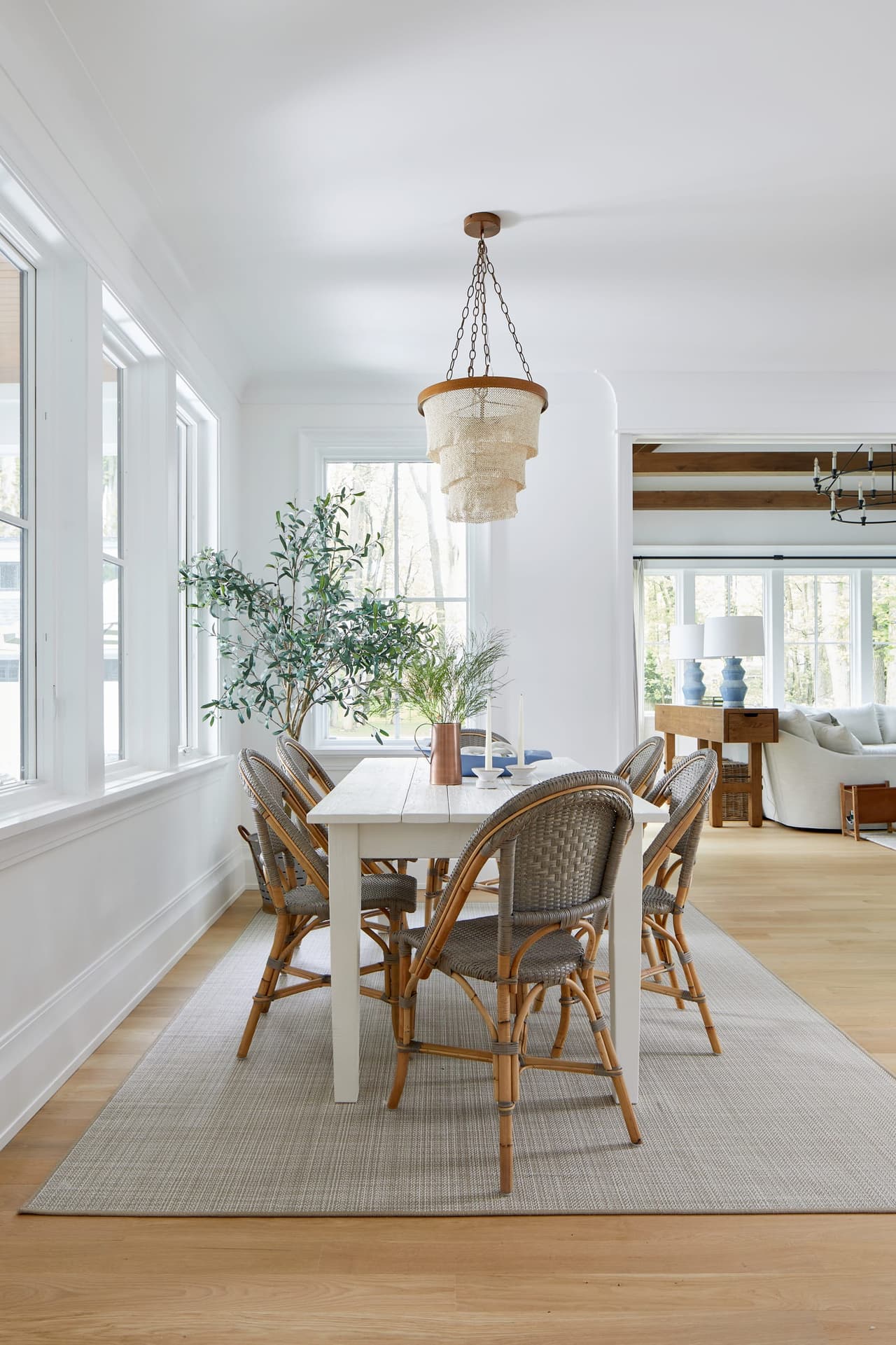 Dining area with a white trestle table, rattan bistro chairs, a woven pendant and an olive tree by the windows