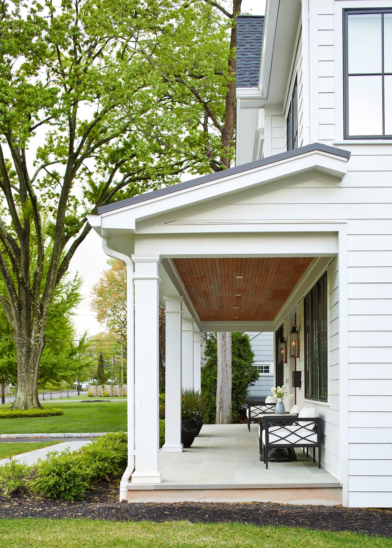 Covered porch of a Westfield, NJ new-construction home seen from the lawn, with a stained plank ceiling, white columns and a bistro table in the shade