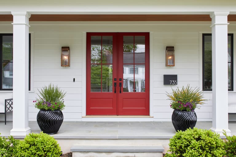 Red double front door between two sconces on the porch of a Westfield, NJ new-construction home, with planted urns and boxwood either side of the steps
