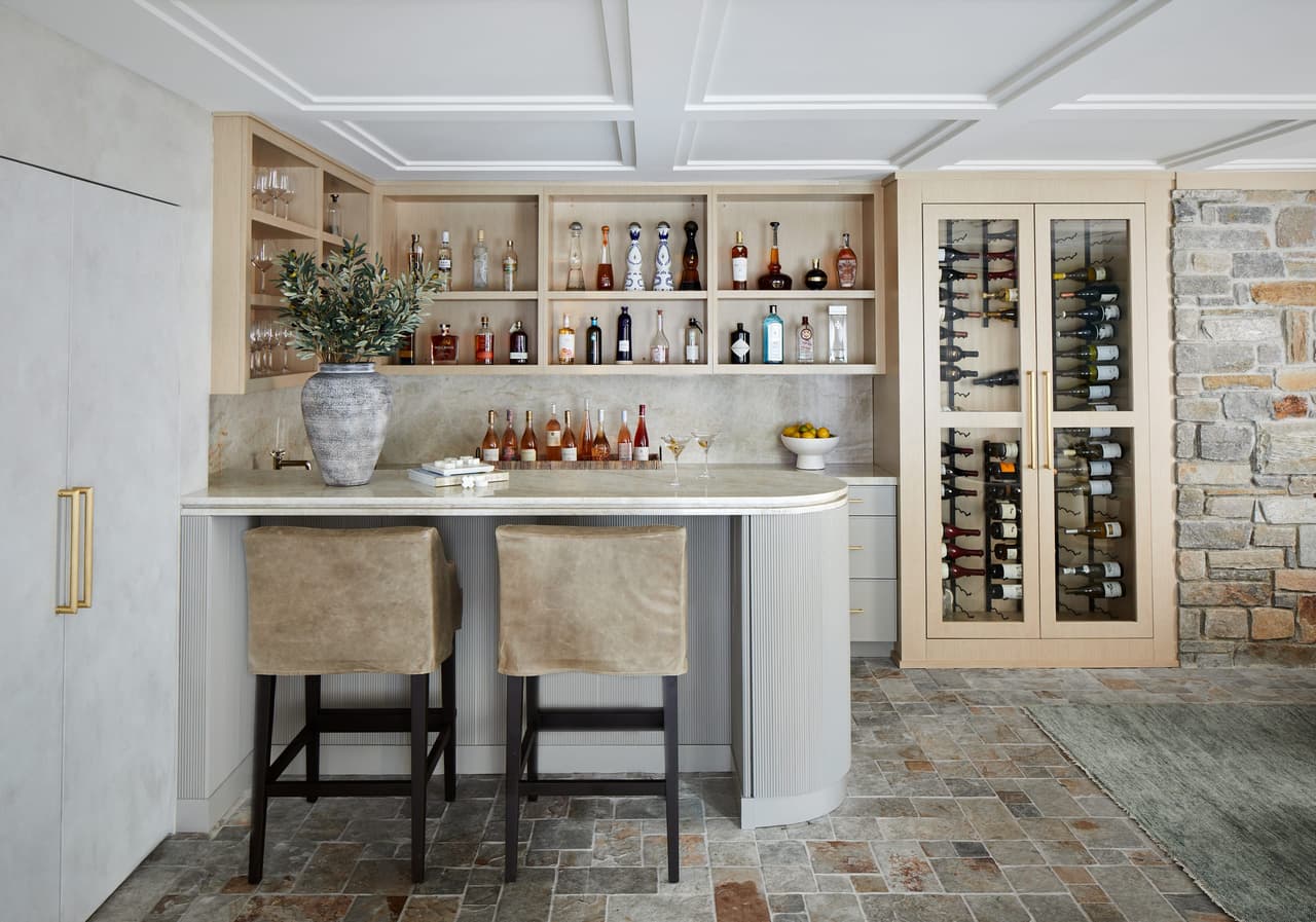 Family room bar in a Westfield, NJ renovation, with a coffered ceiling, open shelving, glass-front wine storage and a stone-clad counter with black stools