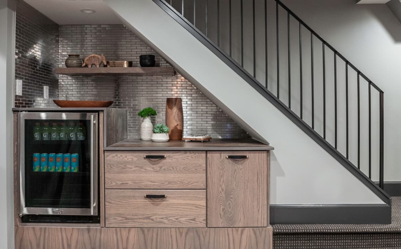 Basement bar built under the stair in a Westfield, NJ renovation, with oak cabinetry, open shelves, a glass-door beverage fridge and a black metal stair rail above