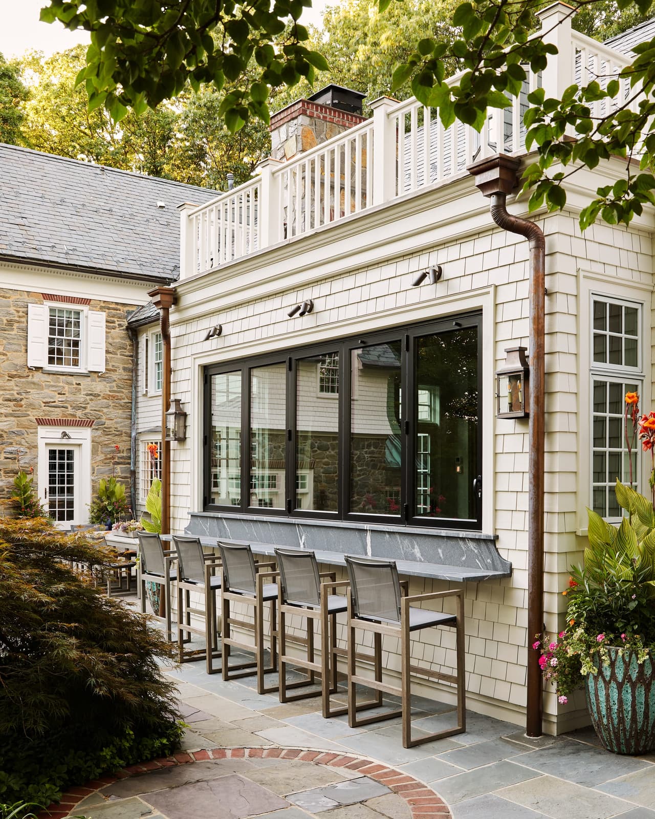 Outdoor bar counter and stools set under the pass-through windows of a shingle and stone Westfield, NJ home renovation, with a copper leader on the wall above