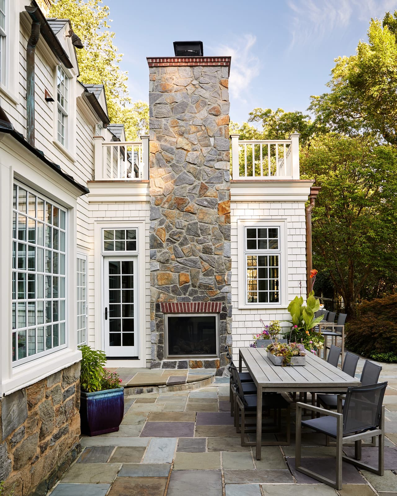 Stone outdoor fireplace rising the full height of a shingle and stone Westfield, NJ home renovation, with a bluestone terrace and dining table below
