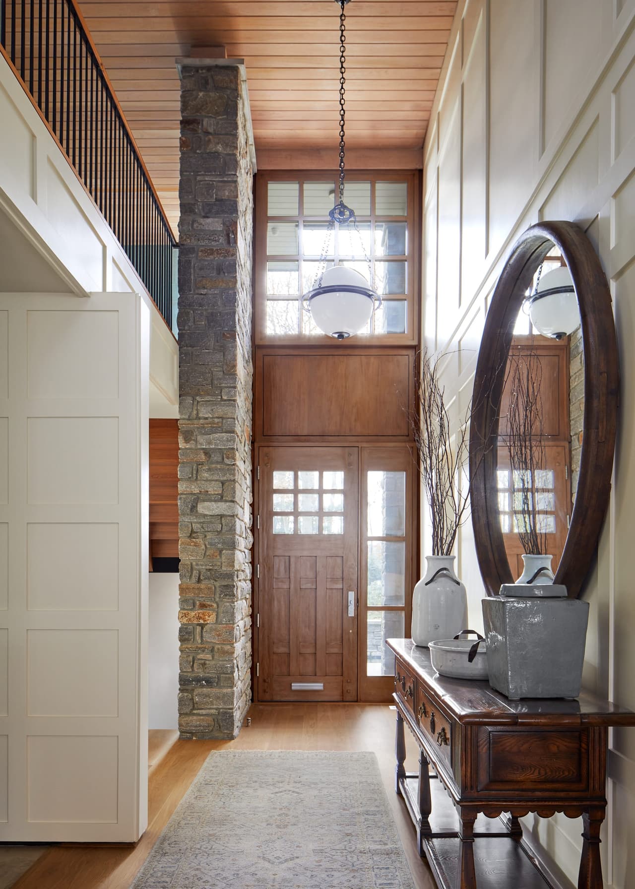 Double-height foyer of a Westfield, NJ renovation with a stone-clad pier, an oversized round mirror and a wood front door with sidelights under a planked ceiling