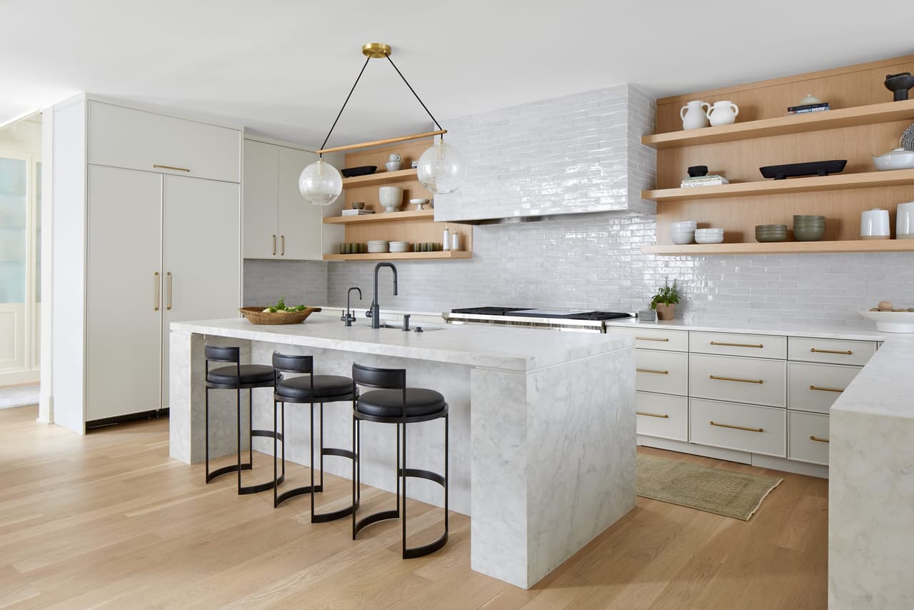 Kitchen of a Westfield, NJ home renovation seen across the marble waterfall island, with custom cabinetry, open shelving either side of the range and a white brick backsplash