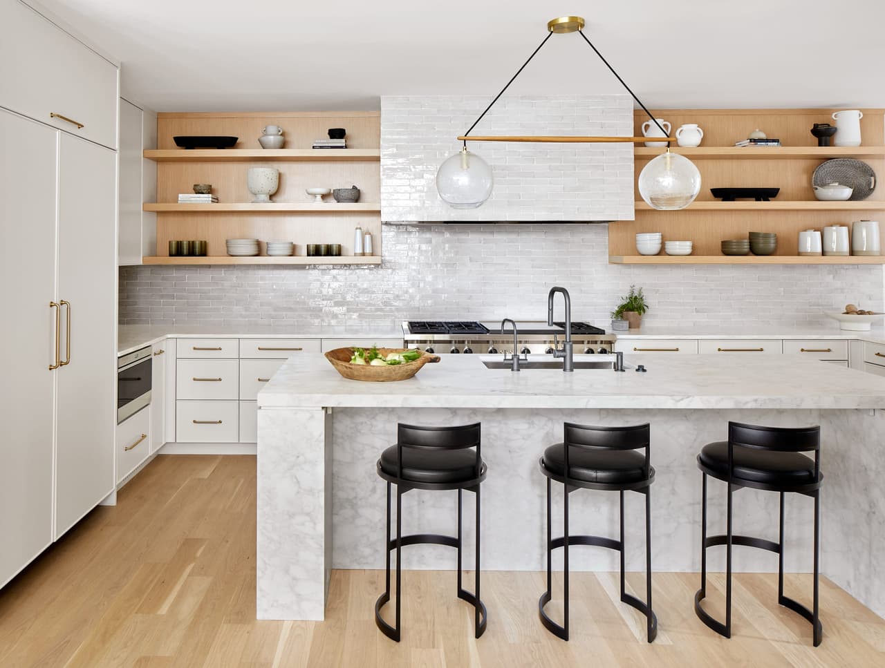 Kitchen island and open oak shelving in a Westfield, NJ home renovation, with a marble waterfall counter, black counter stools and a tiled range wall