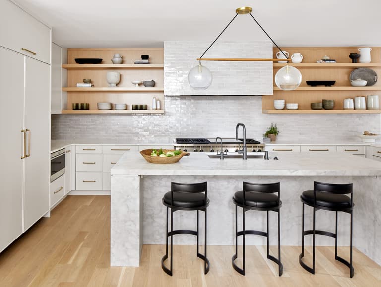 Kitchen island and open oak shelving in a Westfield, NJ home renovation, with a marble waterfall counter, black counter stools and a tiled range wall