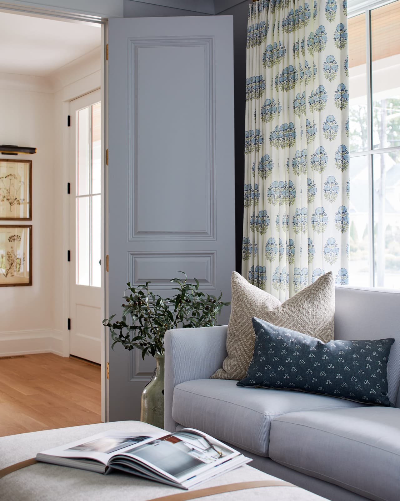Living room of a Westfield, NJ new-construction home with pale blue doors, block-print curtains, a gray sofa with indigo pillows and an olive tree in the corner