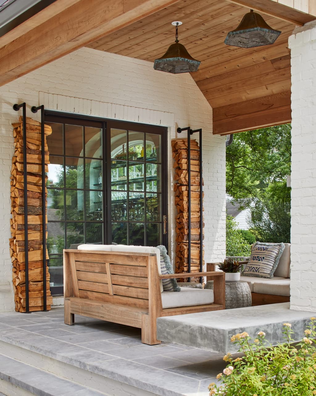 Covered porch of a Westfield, NJ new-construction home by Michael Robert Construction, with a cedar plank ceiling, built-in firewood stacks framing the opening and a hanging daybed
