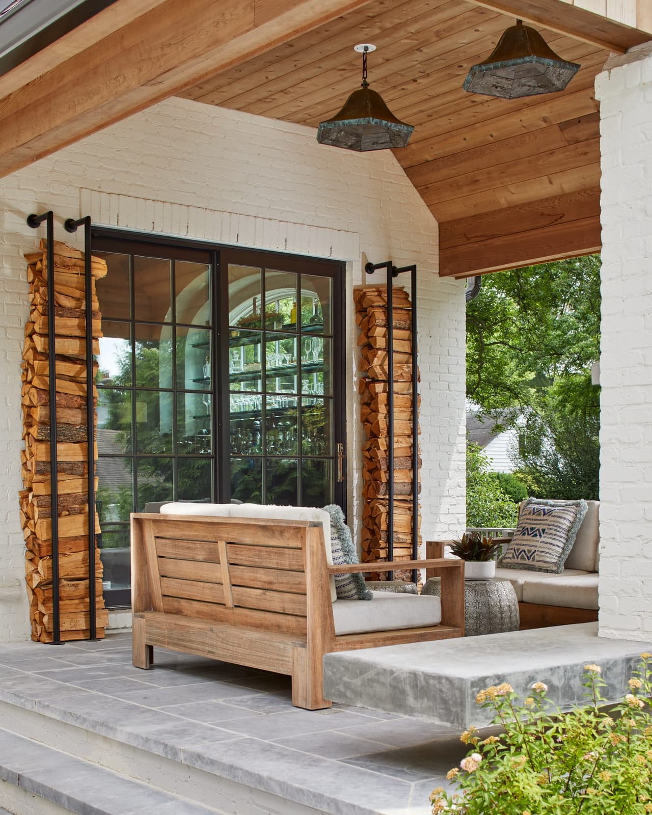 Covered porch of a Westfield, NJ new-construction home with a cedar plank ceiling, built-in firewood stacks framing the opening and a hanging daybed facing the glass doors, built by Michael Robert Construction