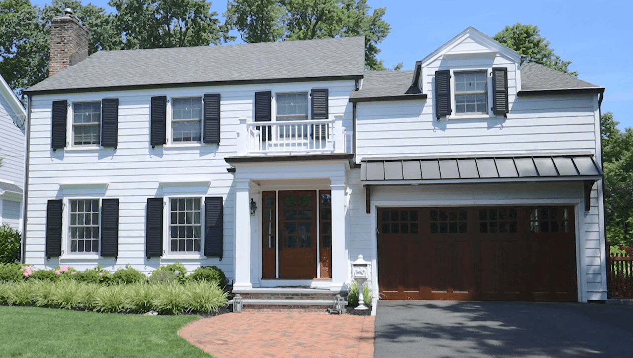 Front of a pale blue Westfield, NJ remodel and addition, with black shutters, a standing-seam porch roof and a wood garage door, built by Michael Robert Construction