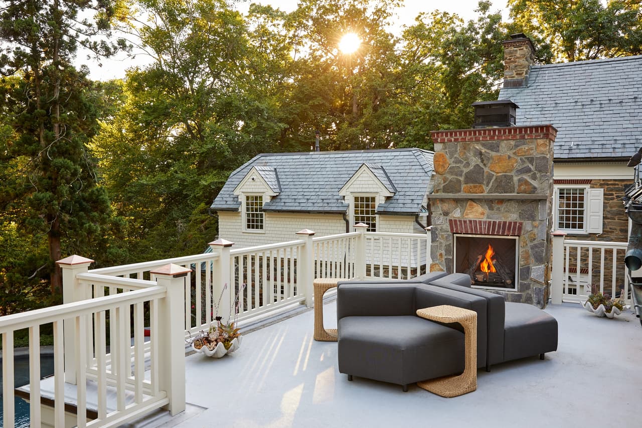Rooftop terrace at a Westfield, NJ stone and shingle renovation, with a lit stone fireplace, a railing in white balusters and the sun through the trees, built by Michael Robert Construction