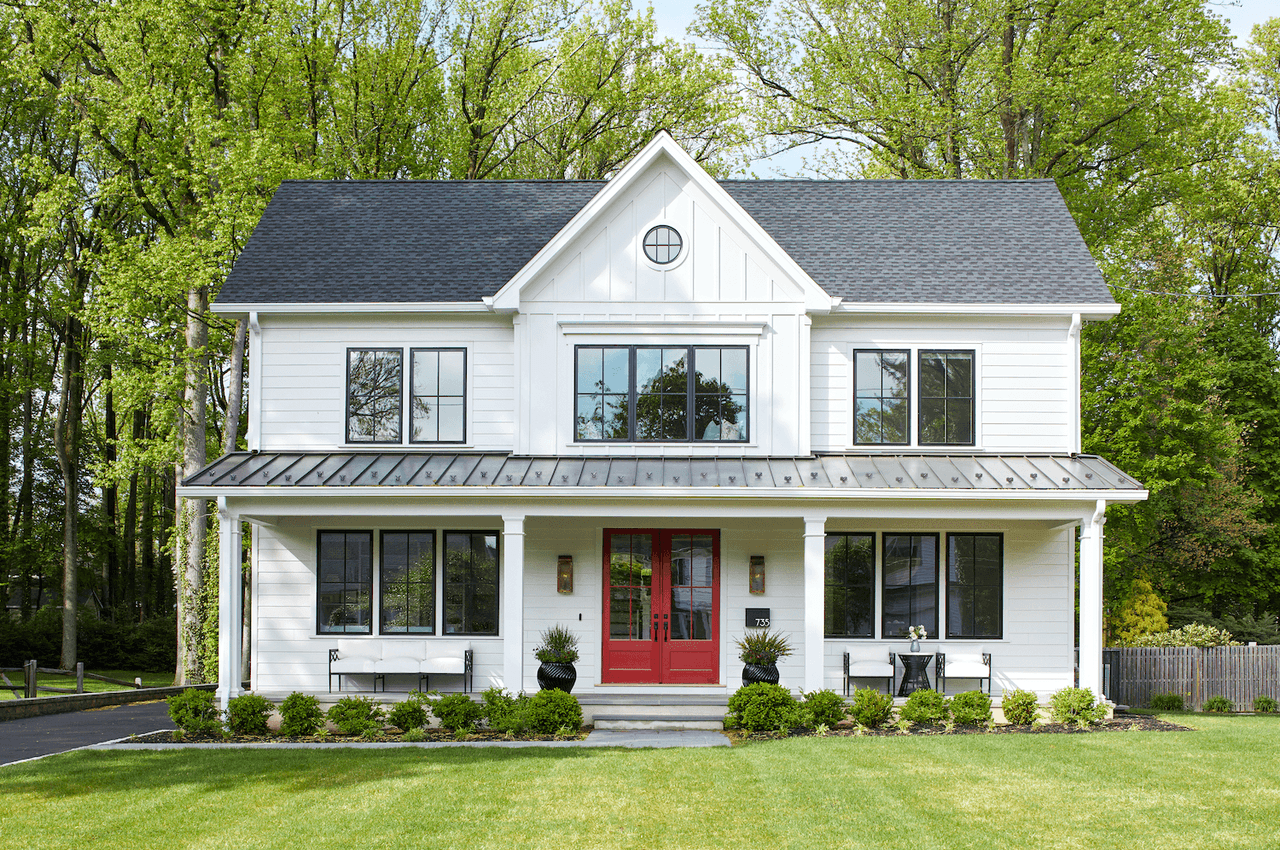 Front elevation of a white modern farmhouse with a red front door, covered porch, gabled dormer and round gable window in Westfield, NJ