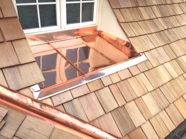 Copper pan flashing installed at the base of a window where it meets cedar shingle siding, the seam soldered and turned up behind the trim