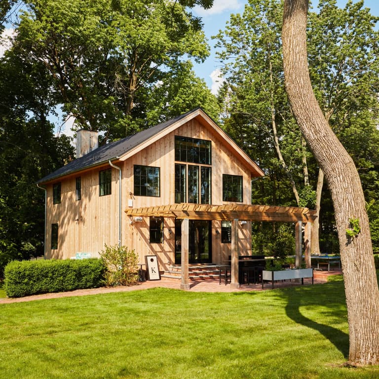 Rear exterior of a cedar-clad barn renovation in Westfield, NJ, with a steep gable, black-framed windows and a pergola over the brick patio, by Michael Robert Construction