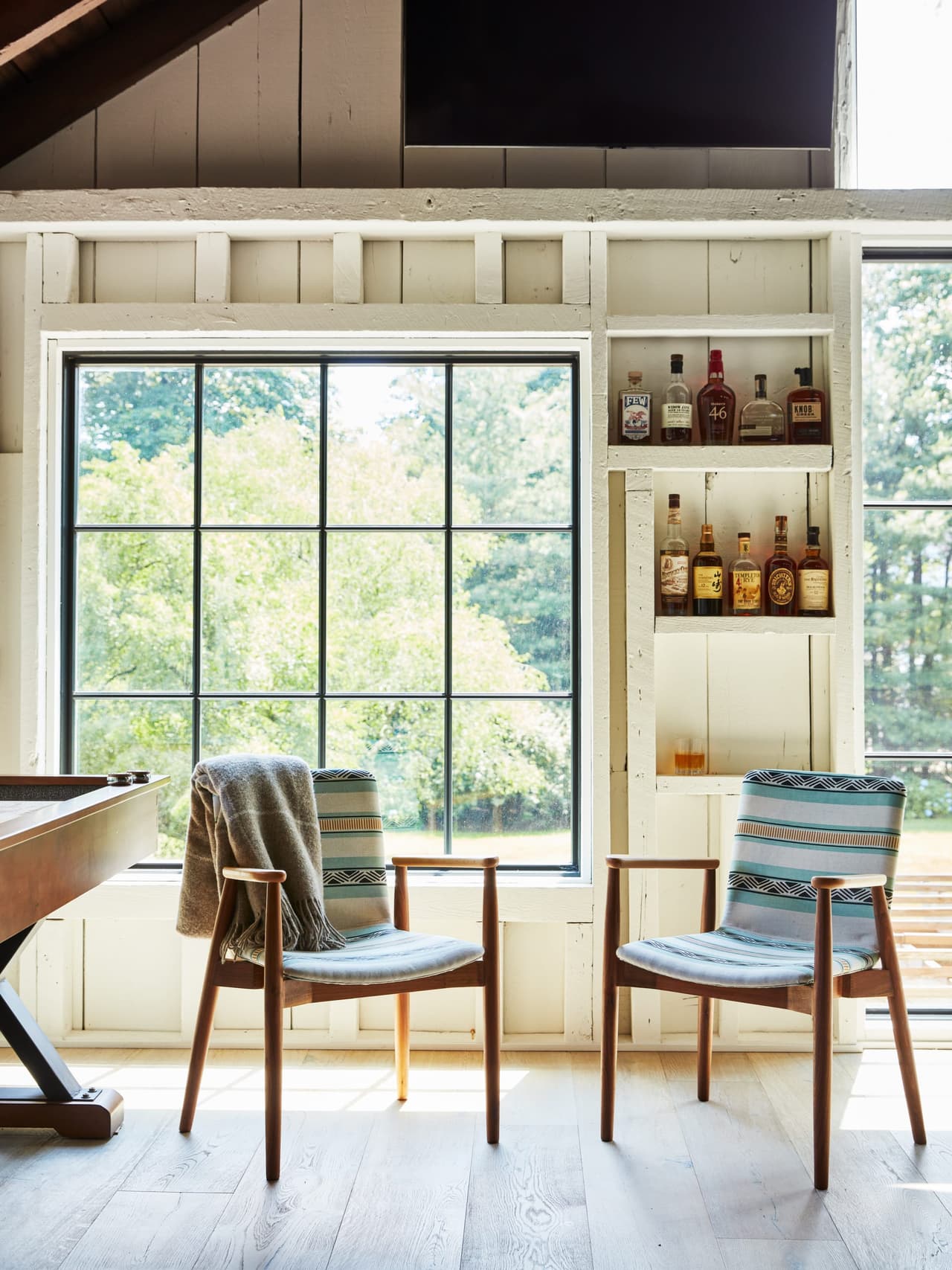 Two chairs at the window of the bar area in a Westfield, NJ barn renovation, with bottle shelves built into the paneling either side
