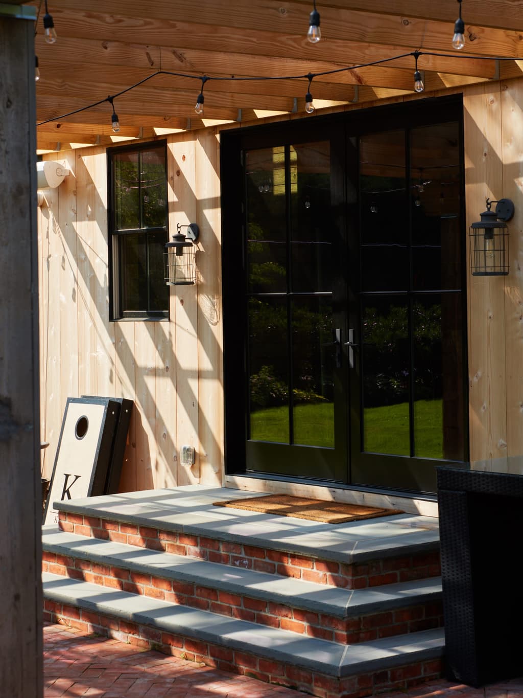 Entry to a historic backyard barn in Westfield, NJ, with black steel-and-glass double doors, a cedar-lined ceiling, string lights and brick-and-slate steps