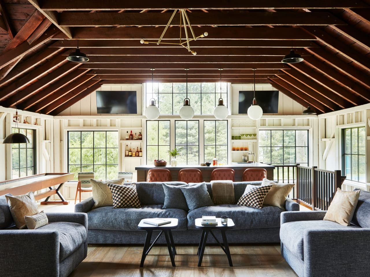 Open-concept family room in a historic backyard barn renovation in Westfield, NJ, under exposed rafters and collar ties, with sectionals facing a wall of black-framed windows