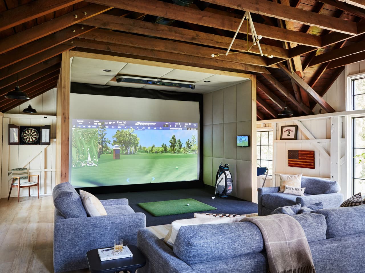 Golf simulator screen set into the barn wall, with sectional seating and a putting strip in front of it