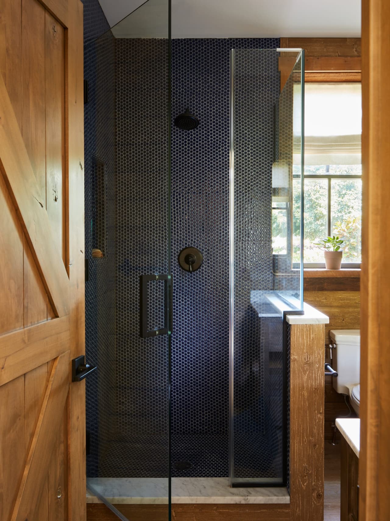Walk-in shower lined in dark penny tile behind glass in a Westfield, NJ barn renovation, beside a plank door and reclaimed wood walls
