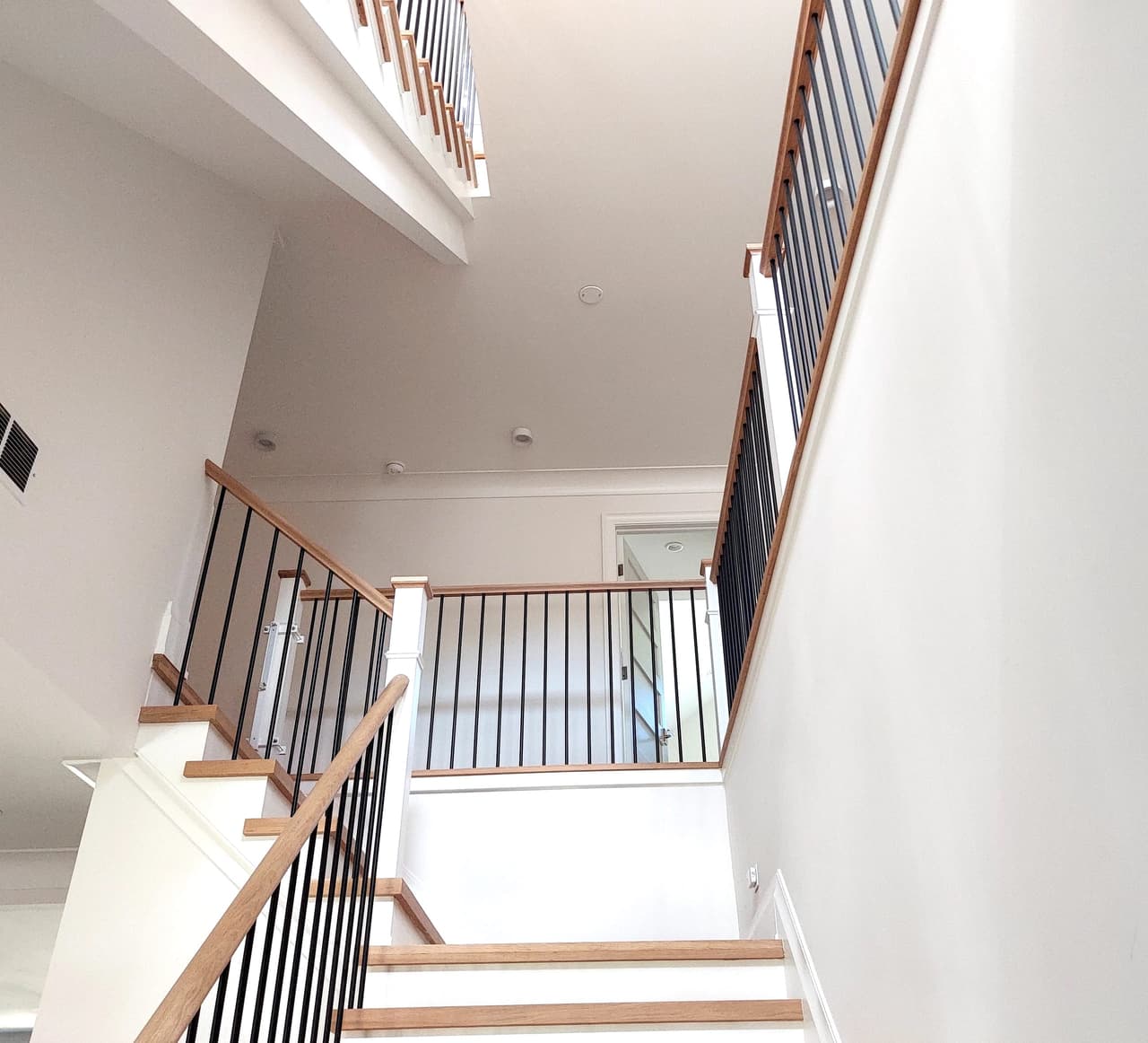 Stair of a Westfield, NJ new-construction home seen from below, with a wood handrail, black metal balusters and daylight from the landing window