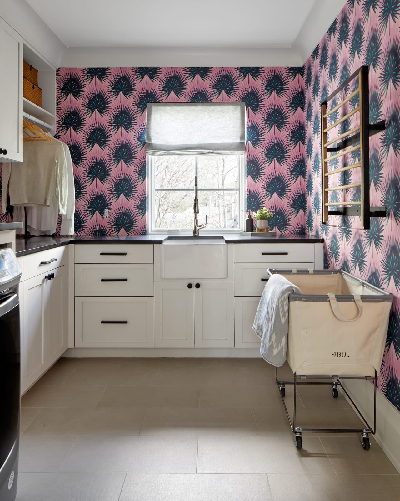 Laundry room in a Westfield, NJ addition and renovation, papered in a pink and navy palm print, with white cabinetry and a drying rail across the window wall