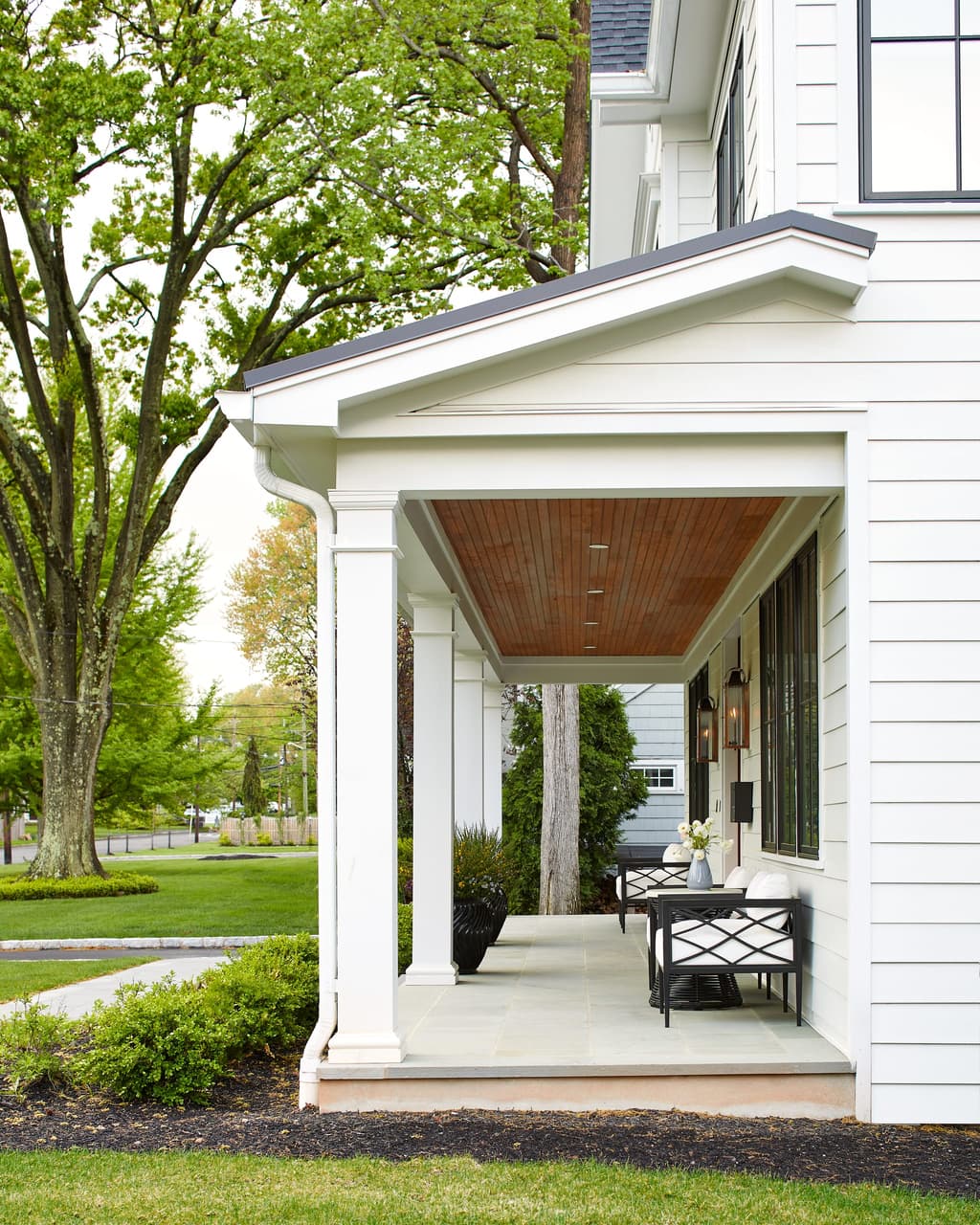Side porch of a Westfield, NJ new-construction modern farmhouse, with a stained plank ceiling, white columns and a bistro table in the shade