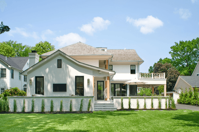 White stucco and shingle home seen across the lawn, with a curved porch roof on wood posts, a covered terrace and arborvitae newly planted along the base