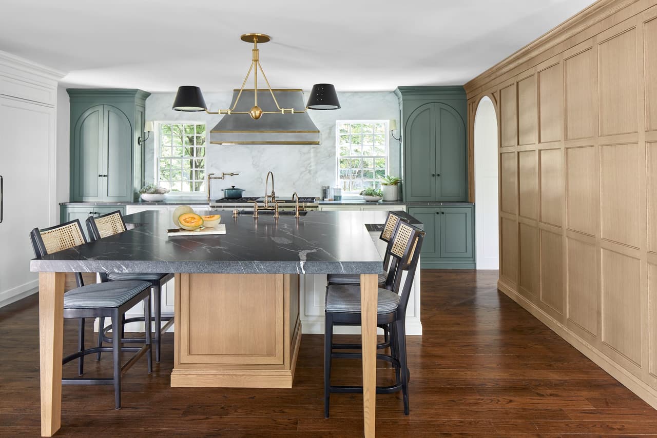 Kitchen of a Westfield, NJ renovation seen along the soapstone island, with sage green cabinetry, a plaster hood and a run of white oak paneling opposite