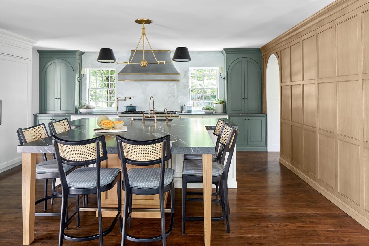 Oak island with spindle stools in a Westfield, NJ kitchen renovation, between sage green cabinetry and a run of white oak paneling with an arched opening