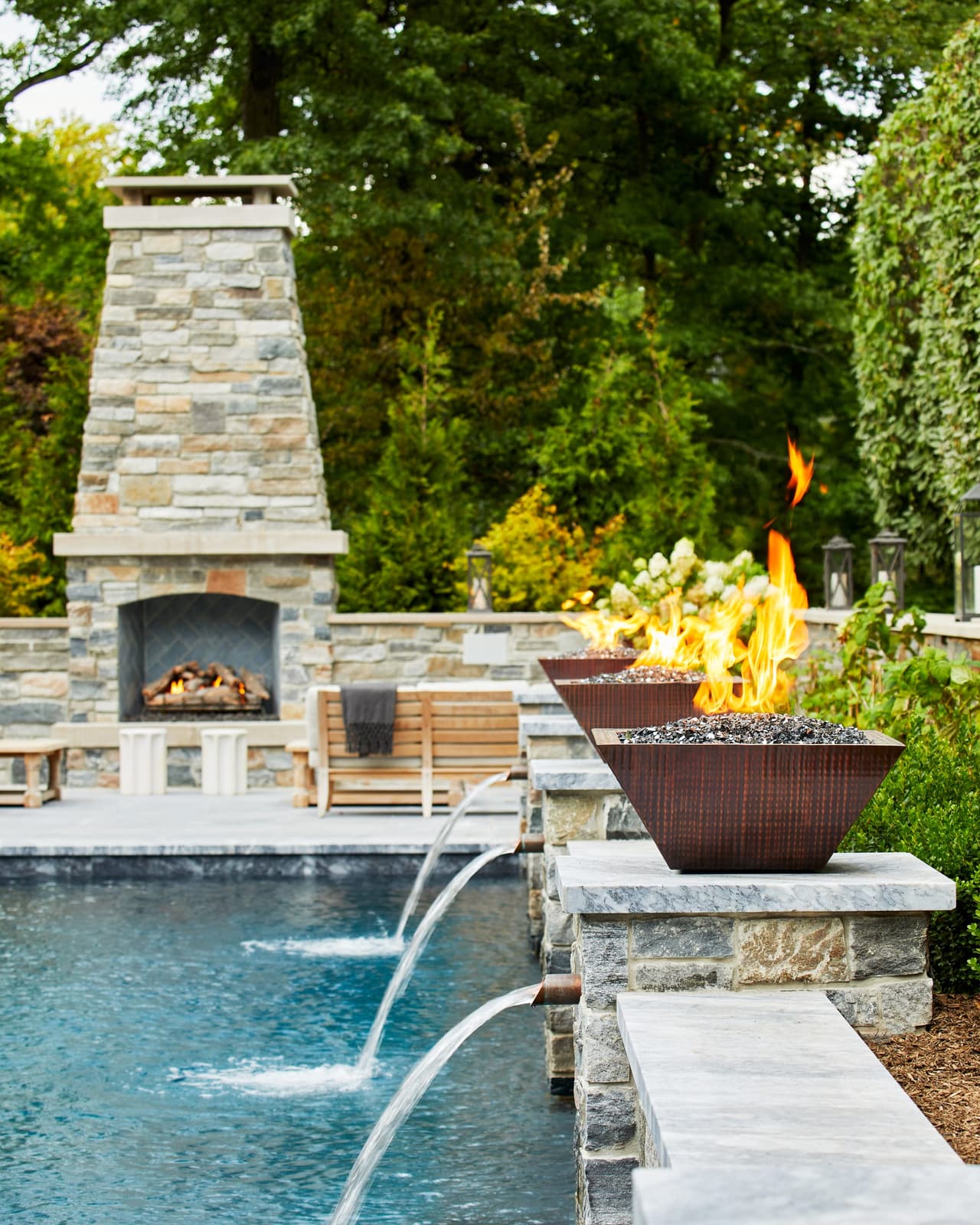 Fire bowl burning above water spouts arcing into the pool of a Westfield, NJ backyard, with a tall stone outdoor fireplace behind, built by Michael Robert Construction