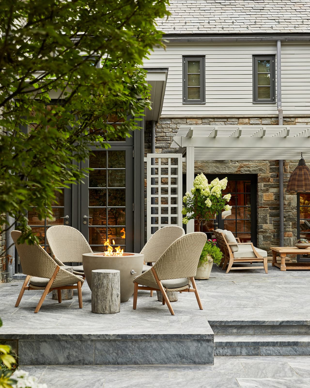 Fire table and woven lounge chairs on the bluestone terrace of a Westfield, NJ home, under a pergola against the stone and shingled rear elevation