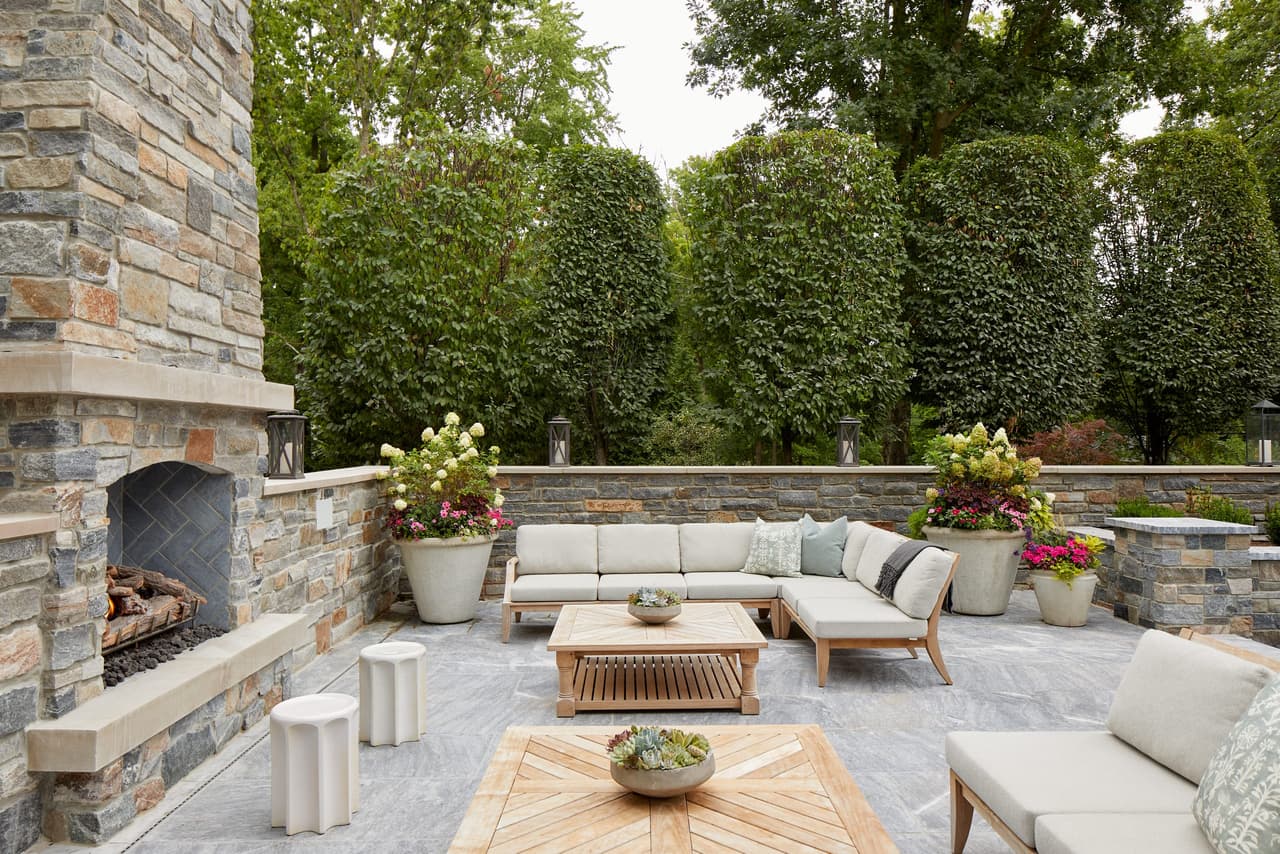 Stone outdoor fireplace and teak sectional on the bluestone terrace of a Westfield, NJ backyard, enclosed by tall clipped hedges